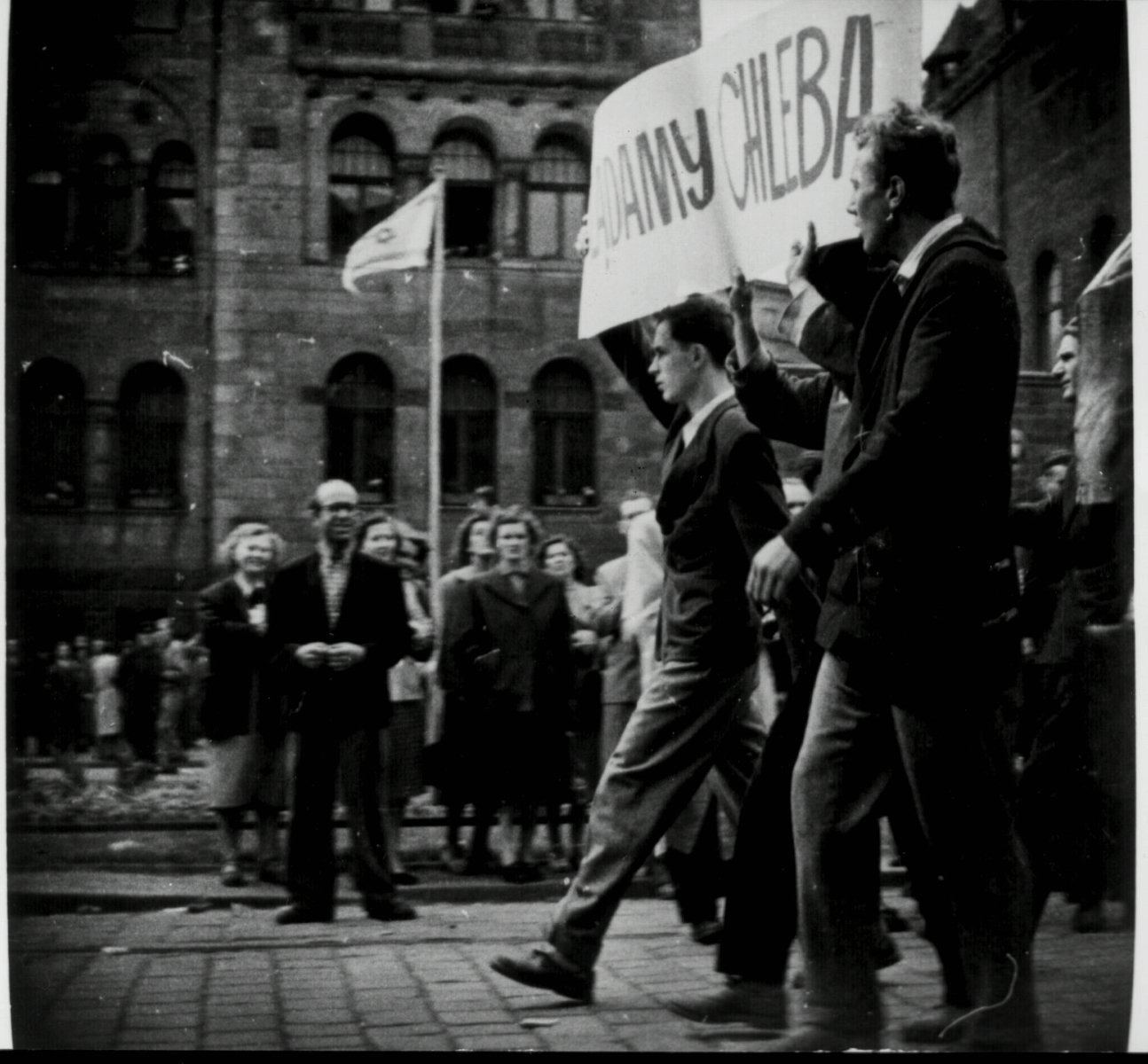 Widok z boku na pochód robotników. Na czele Janusz Kulas (pierwszy) z innymi dwoma mężczyznami niesie transparent "Żądamy chleba". - grafika artykułu