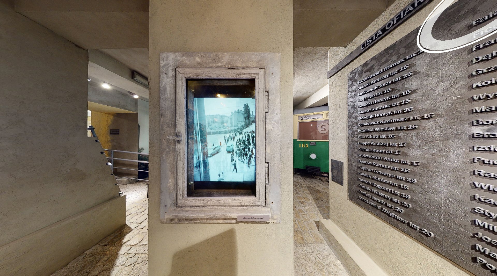 Element wystawy muzealnej, który przedstawia okno Paprzyckiego. W ramie okna kamienicy jest powieszon monitor, który wyświetla zdjęcia Leszka Paprzyckiego. - grafika artykułu