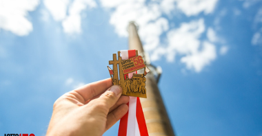 Dłoń trzyma medal z okazji Biegu Pamięci Poznańskiego Czerwca '56. W tle wysoki komin hutniczy, niebieskie niebo i chmury.